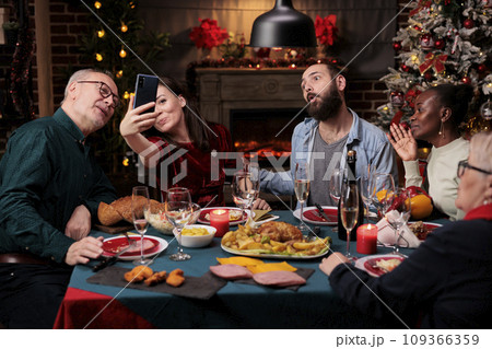 People taking pictures at festive dinner with homemade food and glasses of wine, making memories during christmas eve winter holiday. Diverse persons having fun with photos on phone at home. 109366359