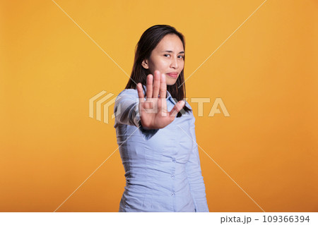 Filipino young adult doing no sign with palm on camera, expressing disapproval and denial. Cheerful woman showing rejection and refusal gesture standing in studio over yellow background Filipino young adult doing no sign with palm on camera, expressing disapproval and denial. Cheerful woman showing rejection and refusal gesture standing in studio over yellow background 109366394