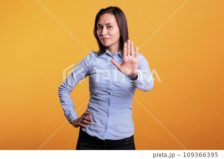 Serious filipino woman doing rejection gesture with hand in front of camera, posing in studio over yellow background. Negative model showing stop gesture, being in denial and expressing refusal. Serious filipino woman doing rejection gesture with hand in front of camera, posing in studio over yellow background. Negative model showing stop gesture, being in denial and expressing refusal. 109366395