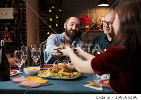 Persons eating food at festive dinner, celebrating christmas eve together and passing meal plates around. Joyful group of people enjoying holiday celebration event at home, winter feast. 109366396