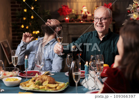Old adult giving toast with raised glass of champagne at christmas eve celebration to enjoy winter holiday with friends and family. Grandpa making speech around the table, sparkling wine. Old adult giving toast with raised glass of champagne at christmas eve celebration to enjoy winter holiday with friends and family. Grandpa making speech around the table, sparkling wine. 109366397