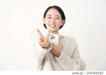 スマホを持ってピースサインをするミドルエイジの女性 白背景 スマホを持ってピースサインをするミドルエイジの女性 白背景 109369382