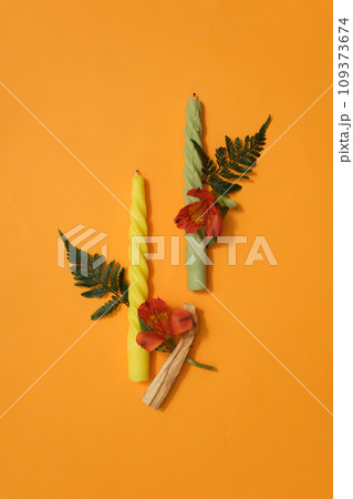 Two twisted candles in two different colors are displayed on a yellow background with fresh flowers and green leaves. Copy space for advertising, magazine posting and more. Two twisted candles in two different colors are displayed on a yellow background with fresh flowers and green leaves. Copy space for advertising, magazine posting and more. 109373674