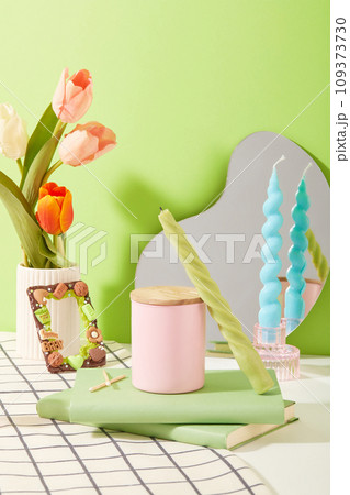 Tulips are placed in a porcelain vase. A jar of scented candles is displayed on the table with two notebooks. Colored candles and a mirror are placed on the table. Mockups for ads. Tulips are placed in a porcelain vase. A jar of scented candles is displayed on the table with two notebooks. Colored candles and a mirror are placed on the table. Mockups for ads. 109373730