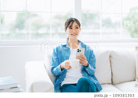 ソファに座ってコーヒーを飲みながらスマホを見る女性 109381303