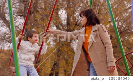 Mother swings boy on swing on park playground. Mother swings preschooler boy on vividly hued swing in park. Mother swings boy in autumn park on brightly colored swing creating memorable family fun Mother swings boy on swing on park playground. Mother swings preschooler boy on vividly hued swing in park. Mother swings boy in autumn park on brightly colored swing creating memorable family fun 109384049