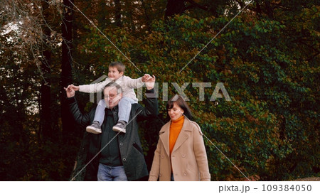 Father and son on shoulders walk in park with accompanying mother. Father and son sitting on shoulders take pleasure walk accompanied by mother. Father and son walk in afternoon with mother in park Father and son on shoulders walk in park with accompanying mother. Father and son sitting on shoulders take pleasure walk accompanied by mother. Father and son walk in afternoon with mother in park 109384050