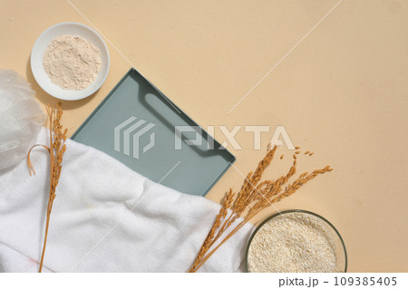 A tray arranged with wheat ears, a towel, a dish of rice bran and a bowl containing rice. Beige background. Vacant space on the tray to show organic products 109385405