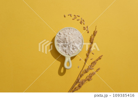 Against yellow background, a ceramic bowl of rice bran is featured with wheat ears. Rice bran in skincare is beloved because it can be used daily Against yellow background, a ceramic bowl of rice bran is featured with wheat ears. Rice bran in skincare is beloved because it can be used daily 109385416