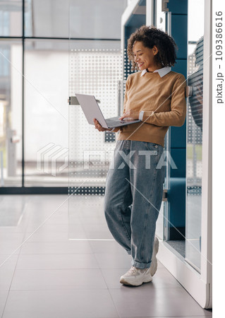 Smiling female freelancer working on laptop while standing on modern coworking background Smiling female freelancer working on laptop while standing on modern coworking background 109386616