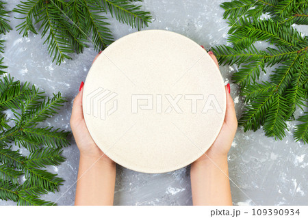 female hands holding an empty white plate on a gray background with Christmas branches. view from above for your design 109390934