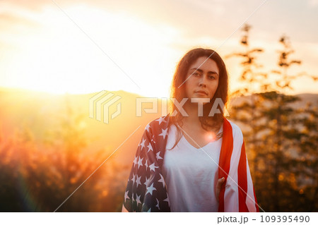 Independence day and Memorial Day. Portrait of patriotic young woman with American flag at shoulders. Sunset in the background. Copy space. The concept of American National Holidays 109395490