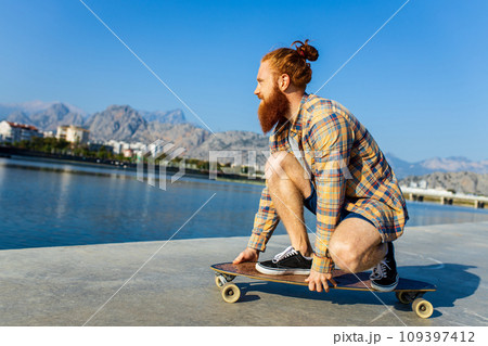 handsome redhaired man with long beard skateboarding near river at sunny summer day handsome redhaired man with long beard skateboarding near river at sunny summer day 109397412