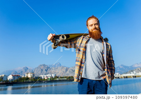 handsome redhaired man with long beard skateboarding near river at sunny summer day 109397480