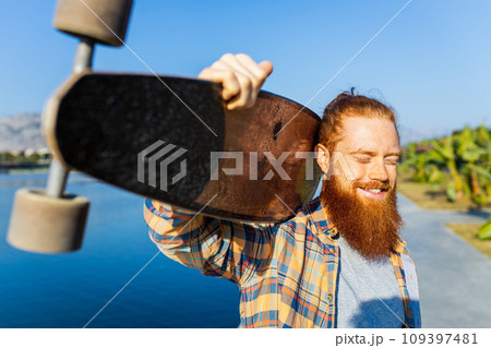 handsome redhaired man with long beard skateboarding near river at sunny summer day 109397481