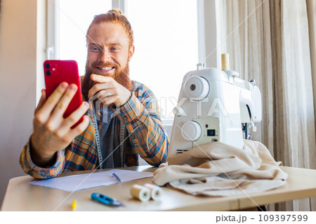 handsome redhaired man with long beard sews at a sewing machine at home studio 109397599