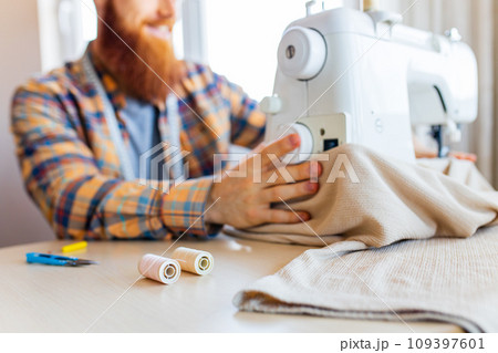 handsome redhaired man with long beard sews at a sewing machine at home studio 109397601
