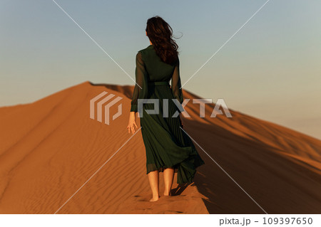 woman in green long dress stands in desert sand dunes background yellow orange sunset 109397650