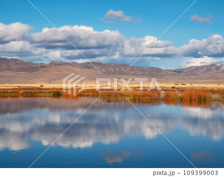 Beautiful autumn mountain lake and mountains. Reflection of snowy peaks and dry yellow grass in the steppe on the surface of a mountain lake. Beautiful autumn mountain lake and mountains. Reflection of snowy peaks and dry yellow grass in the steppe on the surface of a mountain lake. 109399003