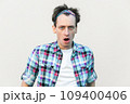 Stupefied man looks with astonishment at camera, being amazed with negative news, does not believe his eyes, wearing checkered shirt and headband. Indoor studio shot isolated on gray background. 109400406