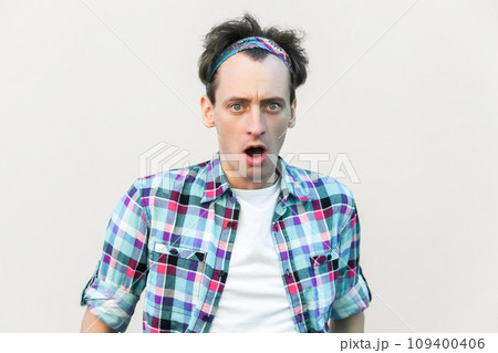 Stupefied man looks with astonishment at camera, being amazed with negative news, does not believe his eyes, wearing checkered shirt and headband. Indoor studio shot isolated on gray background. Stupefied man looks with astonishment at camera, being amazed with negative news, does not believe his eyes, wearing checkered shirt and headband. Indoor studio shot isolated on gray background. 109400406