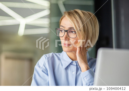 Close-up photo of a young sad businesswoman sitting in the office at a laptop, leaning her head on her hand and looking tiredly to the side. Close-up photo of a young sad businesswoman sitting in the office at a laptop, leaning her head on her hand and looking tiredly to the side. 109400412