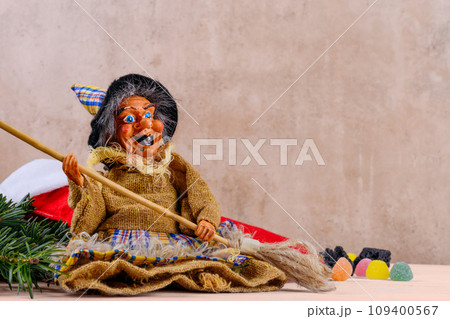 The funny witch Befana and red stocking with sweet coal and candy on beige wall background 109400567