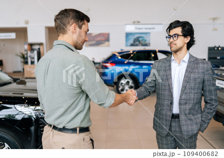 Selective focus of confident car dealer in business suit welcoming young man customer in showroom. Professional salesman shaking client hand in auto dealership. Concept of buying new car. 109400682