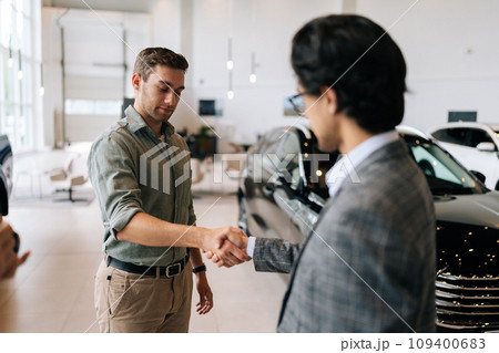 Rear view of unrecognizable car dealer in business suit welcoming successful male customer in showroom. Professional salesman shaking young man client hand. Auto business, trading, service. 109400683