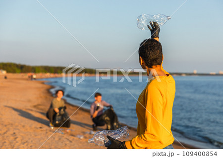 Earth day. Volunteers activists team collects garbage cleaning of beach coastal zone. Woman puts plastic bottle trash in garbage bag on ocean shore. Environmental conservation coastal zone cleaning 109400854