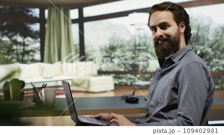 Rich adult seated at desk in his expensive lodge with massive windows displaying breathtaking forest scenery, occupied with contemplating about additional income. Investor in mountaintop villa. 109402981