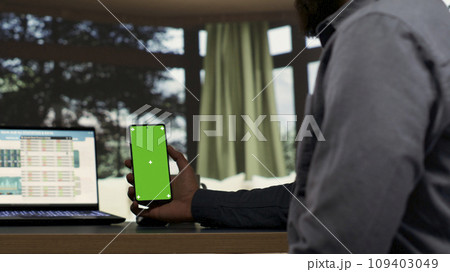 Male billionaire uses greenscreen phone display at desk in his opulent alpine retreat, rich entrepreneur at vacation home. Young shareholder works with isolated chromakey, investing capital funds. 109403049