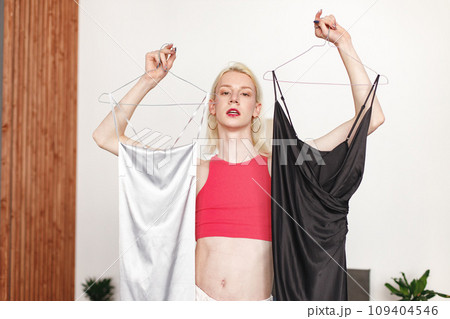 Man with make up on his face standing in a bedroom in his flat. Man wearing fashionable pink top, white trousers. Man choosing what to wear: black or white dress. 109404546