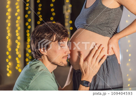 In a tender holiday moment, a husband kisses his wife's pregnant belly, expressing love and anticipation for the Christmas joy to come In a tender holiday moment, a husband kisses his wife's pregnant belly, expressing love and anticipation for the Christmas joy to come 109404593
