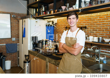 Professional young coffee barista standing inside the counter bar in the coffee shop and making arms crossed and looking at camera, handsome - good looking caucasian ethnic barista posing. Professional young coffee barista standing inside the counter bar in the coffee shop and making arms crossed and looking at camera, handsome - good looking caucasian ethnic barista posing. 109410807