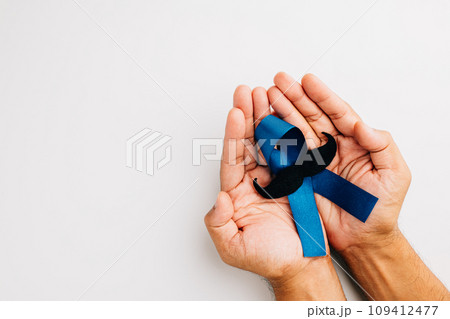 Supporting men living with cancer in November. A man's hands cradle a light blue ribbon with a mustache on a blue background, representing unity and strength. 109412477
