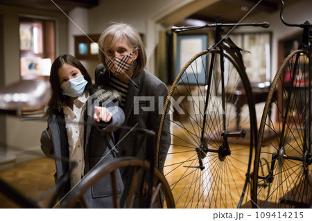 Elderly woman with preteen granddaughter in protective masks examining retro bicycles in museum 109414215