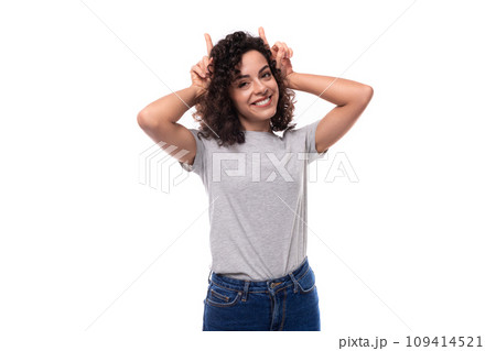 young funny pretty brunette curly caucasian lady in casual jeans makes a face with horns on a white 109414521