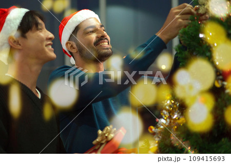 Lovely gay couple enjoy decorating a Christmas tree together in a living room with beautiful festive light bulbs and presents. LGBTQ gay couple celebrating a Christmas Eve and New Year festival. Lovely gay couple enjoy decorating a Christmas tree together in a living room with beautiful festive light bulbs and presents. LGBTQ gay couple celebrating a Christmas Eve and New Year festival. 109415693