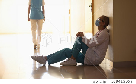 Doctor in medical mask sits on floor in hospital corridor and walking nurse in background Doctor in medical mask sits on floor in hospital corridor and walking nurse in background 109415955