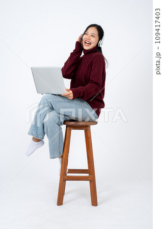 Young happy Asian woman enjoys listening to music through headphones while sitting on a chair. Young happy Asian woman enjoys listening to music through headphones while sitting on a chair. 109417403