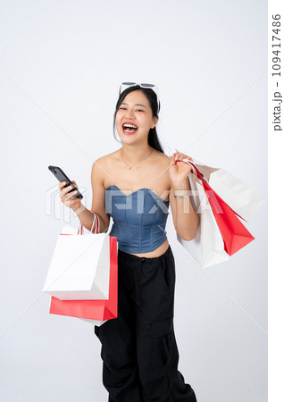 A cheerful and elegant Asian woman in trendy clothes is holding her smartphone and shopping bags. 109417486