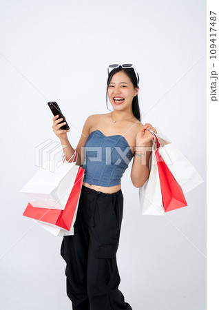 A cheerful and elegant Asian woman in trendy clothes is holding her smartphone and shopping bags. 109417487