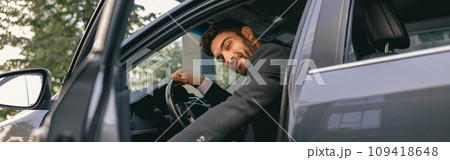 Smiling businessman in suit looking at camera while sitting in driver seat with opened door Smiling businessman in suit looking at camera while sitting in driver seat with opened door 109418648
