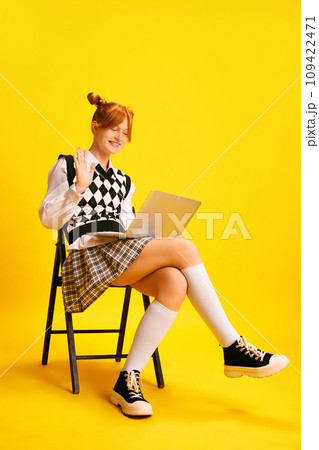 Happy cheerful girl sitting on chair and waving hello to laptop camera as she studies, holds conference online against yellow background. Happy cheerful girl sitting on chair and waving hello to laptop camera as she studies, holds conference online against yellow background. 109422471