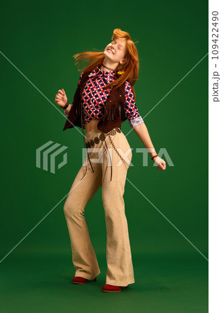 Full length portrait of cheerful young girl, teenager dressed like cowgirl and wind blowing her red hair while she dancing against green studio background. Full length portrait of cheerful young girl, teenager dressed like cowgirl and wind blowing her red hair while she dancing against green studio background. 109422490