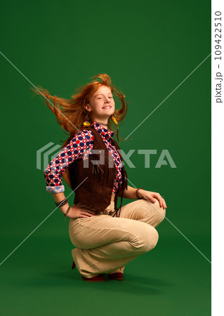 Full length portrait of beautiful red-haired girl, cowgirl sitting and wind blowing her hair against green studio background. 109422510
