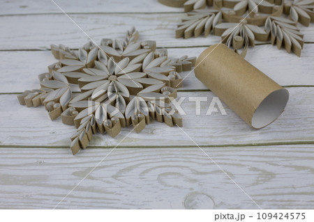 Craft a snowflake from toilet paper rolls. New Year's homemade toys from scrap materials. Home holiday decorations made from recycled materials on white wooden background Craft a snowflake from toilet paper rolls. New Year's homemade toys from scrap materials. Home holiday decorations made from recycled materials on white wooden background 109424575