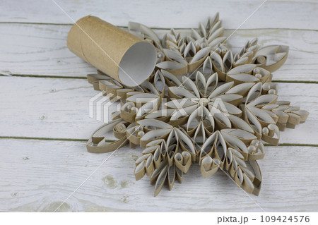 Craft a snowflake from toilet paper rolls. New Year's homemade toys from scrap materials. Home holiday decorations made from recycled materials on white wooden background Craft a snowflake from toilet paper rolls. New Year's homemade toys from scrap materials. Home holiday decorations made from recycled materials on white wooden background 109424576
