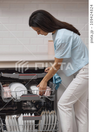 pretty woman loading dishes to open automatic built-in dishwasher machine with clean utensils inside in modern white kitchen. pretty woman loading dishes to open automatic built-in dishwasher machine with clean utensils inside in modern white kitchen. 109424824